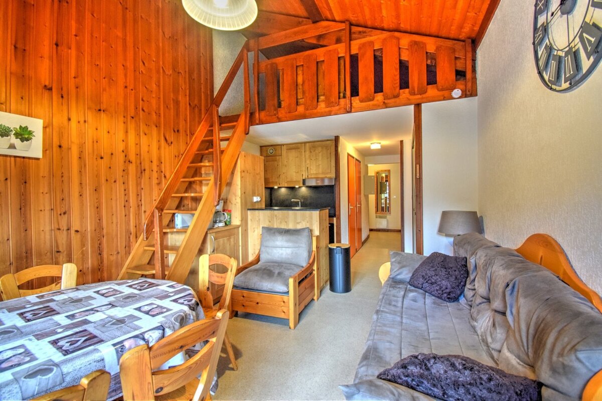A rustic, wood-paneled apartment with a wooden loft, stairs, dining table, kitchenette, and a sofa, offering a cozy chalet atmosphere.