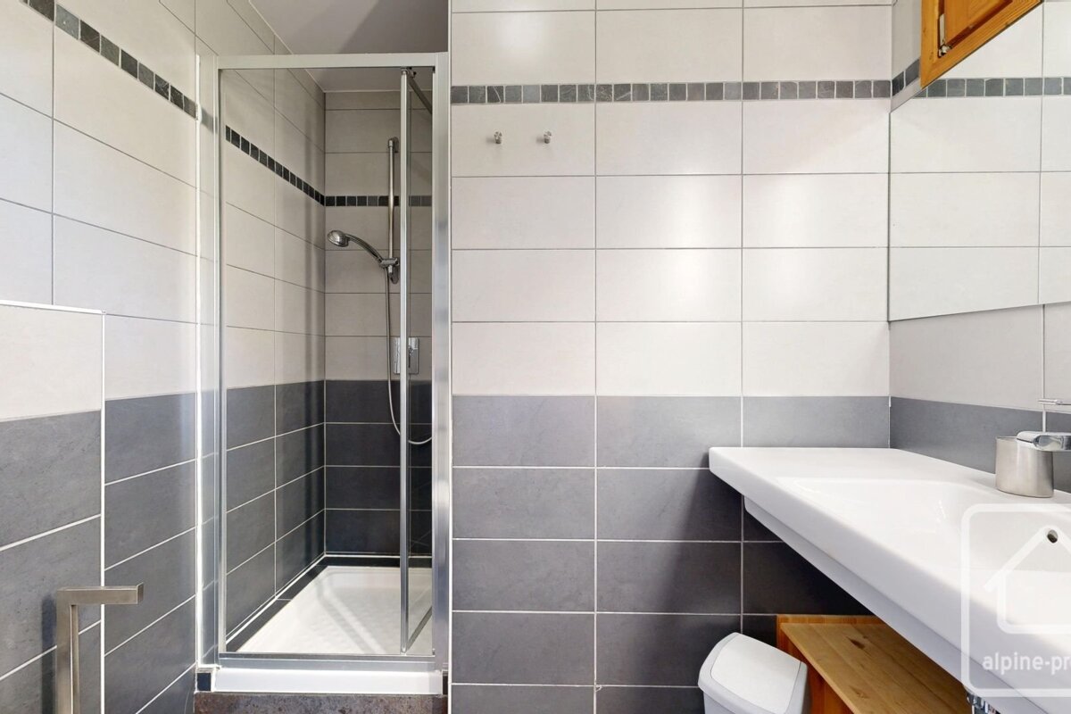 A modern bathroom featuring a walk-in glass shower, white and grey tiled walls, and a sink basin on the right.