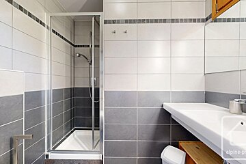 A modern bathroom featuring a walk-in glass shower, white and grey tiled walls, and a sink basin on the right.