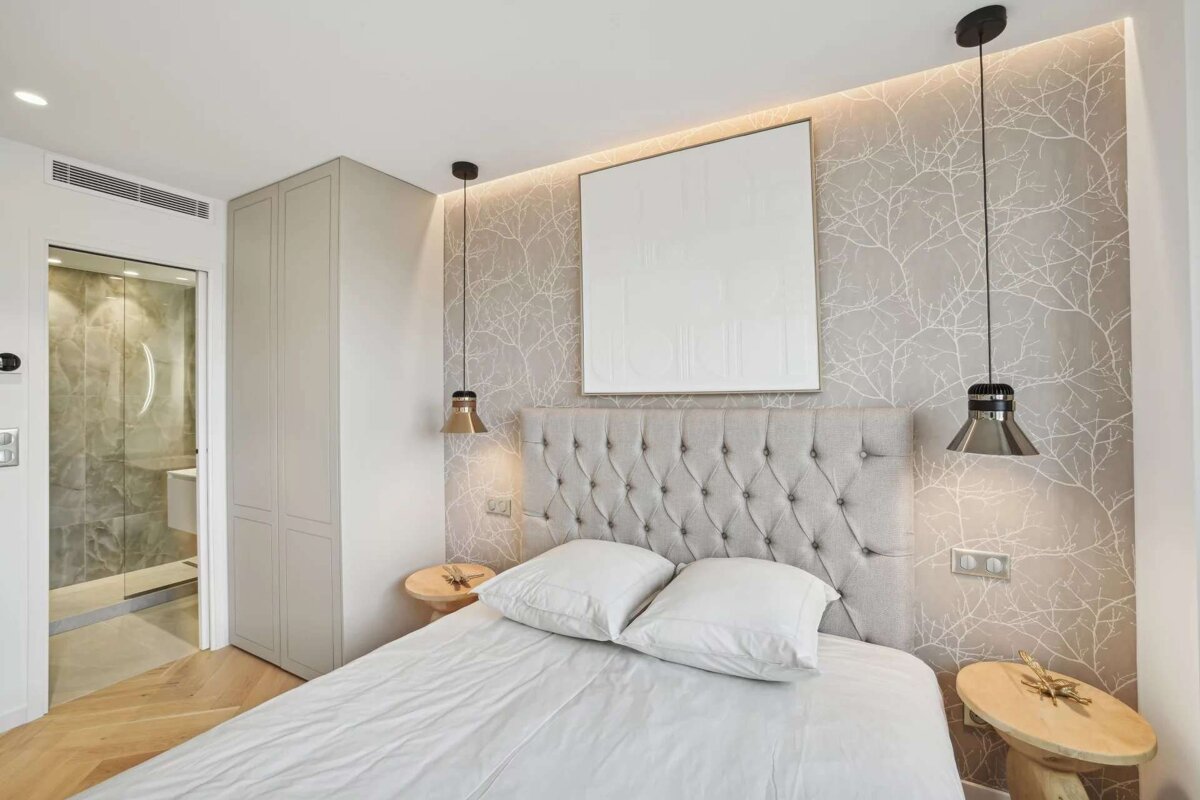 A modern bedroom features a gray tufted headboard, tree branch wallpaper, white bedding, and pendant lights. An adjoining bathroom is visible.