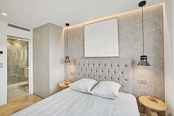 A modern bedroom features a gray tufted headboard, tree branch wallpaper, white bedding, and pendant lights. An adjoining bathroom is visible.
