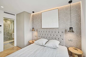 Modern bedroom with a gray tufted headboard, tree branch wallpaper, and pendant lights. White bedding, wooden nightstands, and an en-suite bathroom visible.