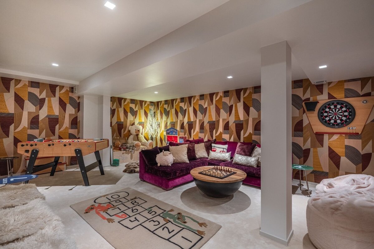 A vibrant game room with abstract wallpaper, featuring a foosball table, dartboard, purple sofa, and a hopscotch rug.
