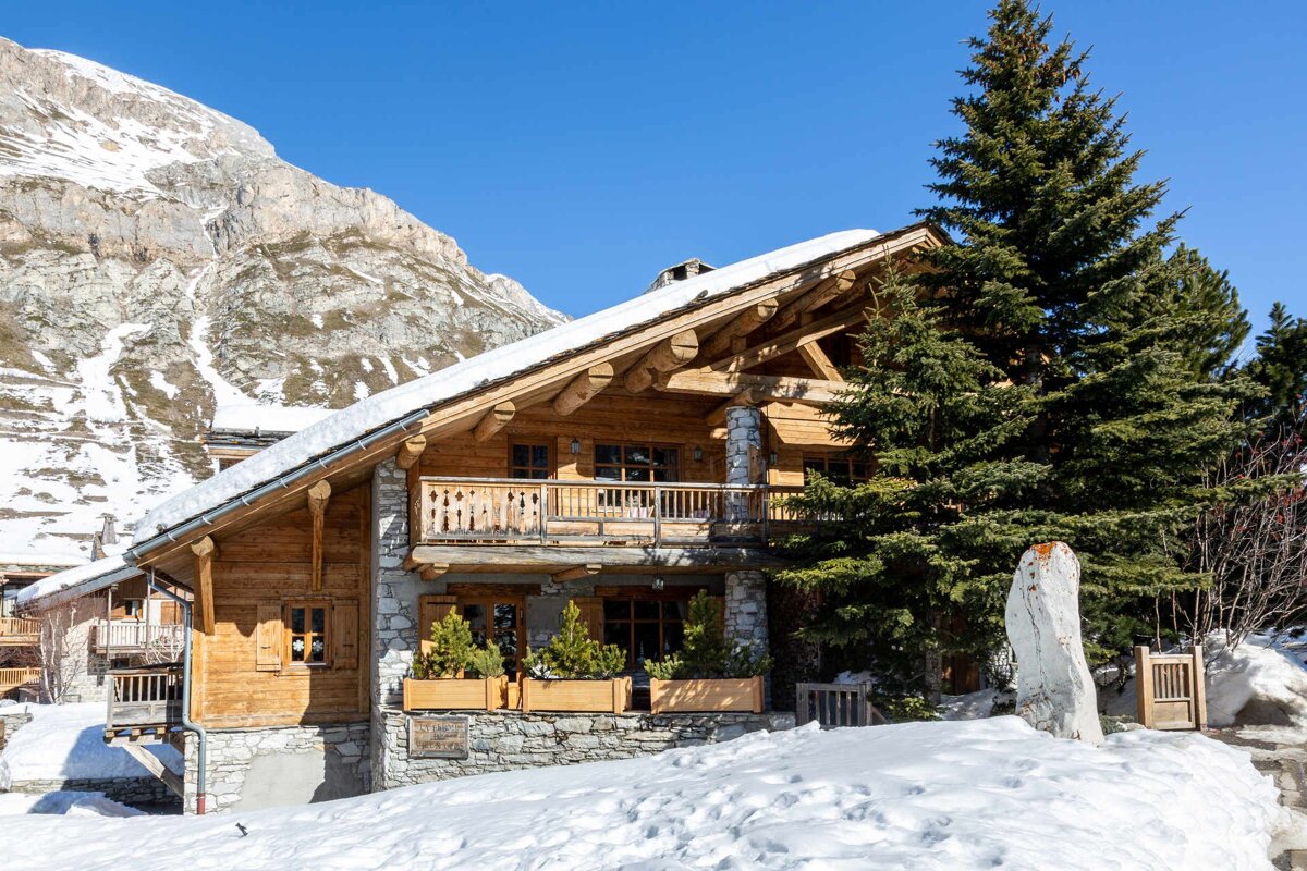 A rustic wooden chalet with stone accents, snow-covered roof, and pine trees, set against a snowy mountain and clear blue sky.