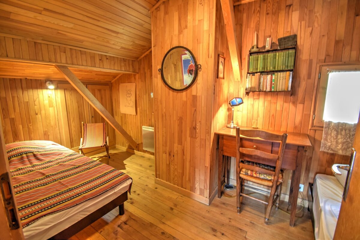 A cozy, rustic room fully paneled in wood with a bed, desk, bookshelf, and mirror. Warm lighting highlights the natural wood aesthetic.