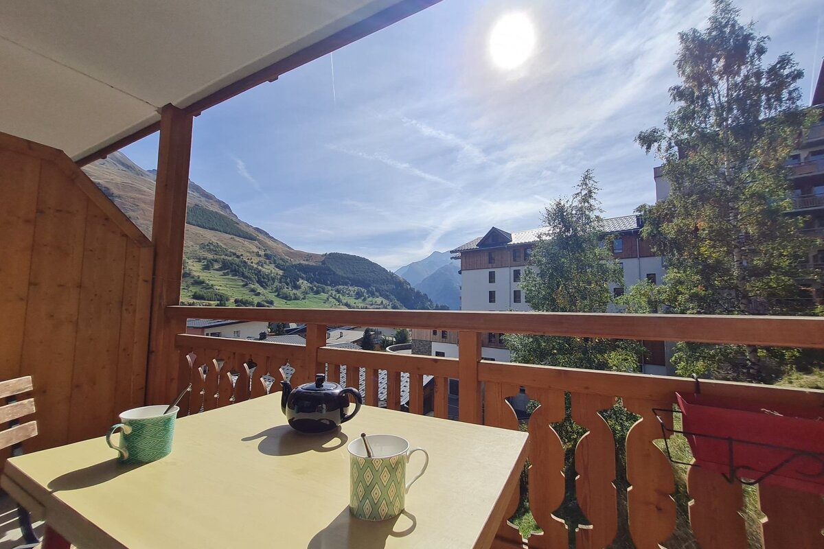 A sunny balcony with a teapot and mugs overlooks a scenic mountain valley, village buildings, and a bright sky with sun.