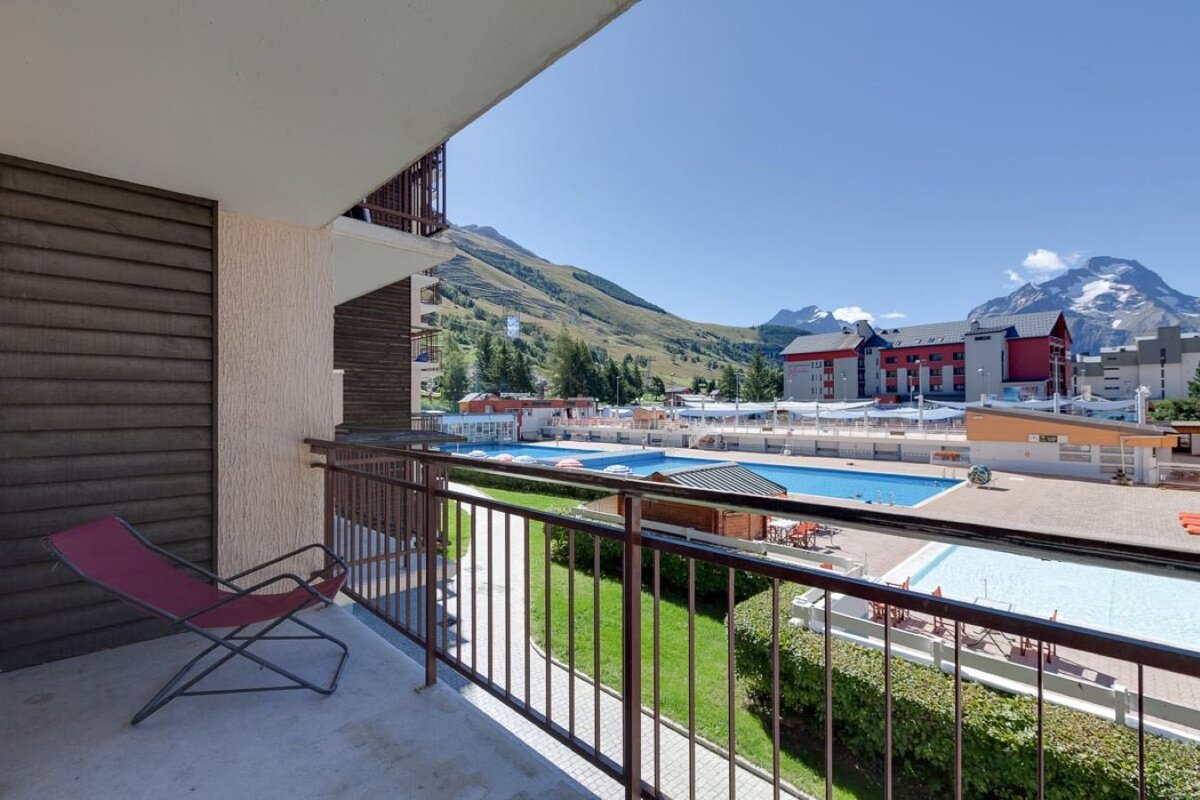 A balcony overlooks a sunny mountain resort with multiple swimming pools, modern buildings, and green hills under a clear sky. A red chair sits on the balcony.