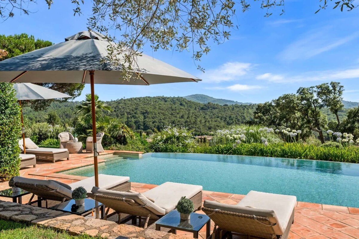 A luxurious outdoor infinity pool area with lounge chairs and umbrellas, set against a backdrop of lush green mountains and a bright blue sky.