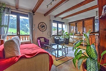 A cozy living room with large windows overlooking a green mountain landscape. Features a red sofa, armchairs, wooden beams, and indoor plants.
