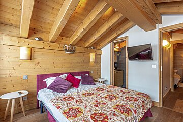 Cozy wooden chalet bedroom featuring exposed ceiling beams, a colorful patterned quilt, and purple pillows.