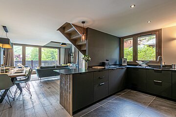 Modern open living area with a sleek dark kitchen, dining space, lounge, and wooden staircase. Large windows offer a lush mountain view.