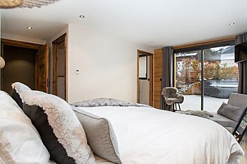 A cozy modern chalet bedroom with plush pillows, wooden accents, and glass doors opening to a snowy outdoor patio.