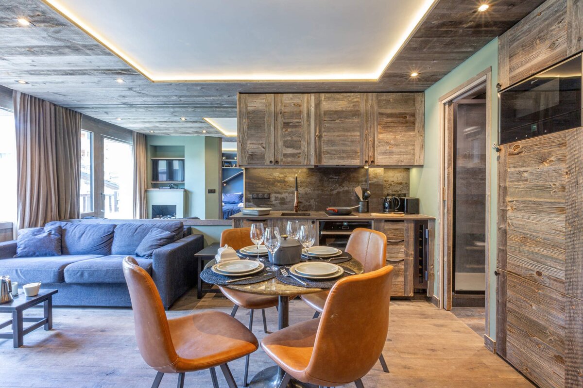 Modern rustic chalet interior featuring a dining table with leather chairs, a blue sofa, and warm wood-paneled walls and ceilings.