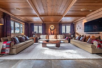 A cozy, wood-paneled chalet-style living room with three beige sofas, a large rug, a unique wooden coffee table, windows, and a big TV.