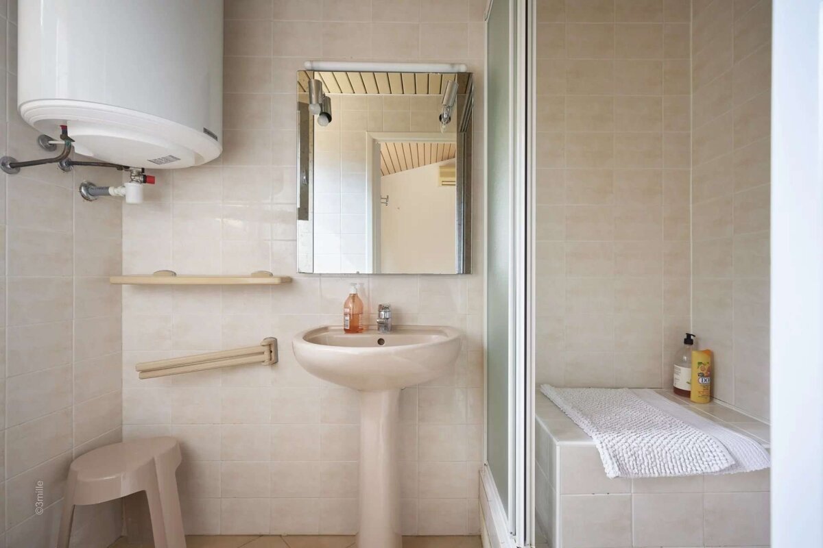 A neutral-toned tiled bathroom with a water heater, pedestal sink, mirror, and a shower stall featuring a tiled bench with a white mat.