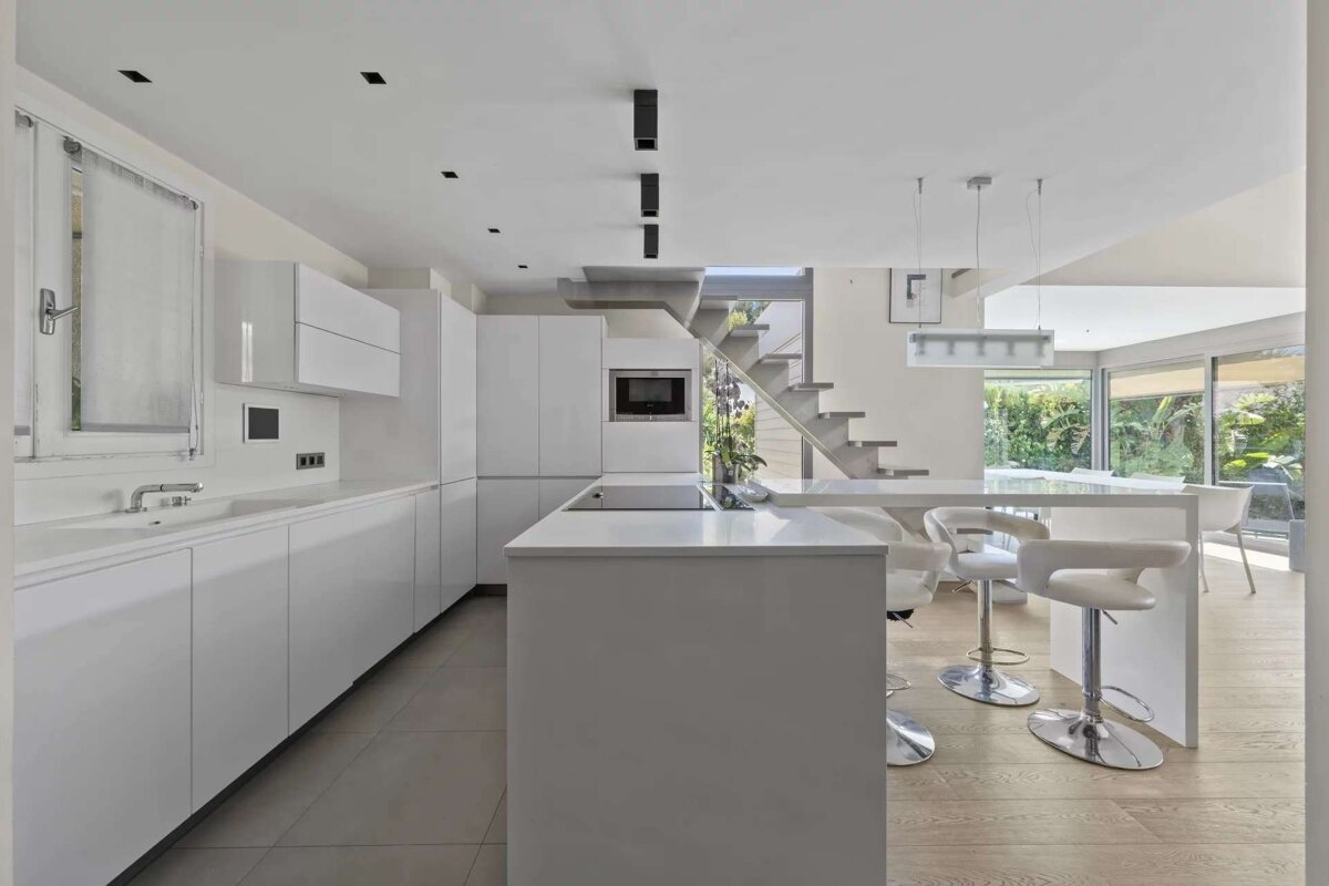 A bright, minimalist white kitchen with an island, bar stools, and modern appliances. An open layout leads to a contemporary staircase and garden views.