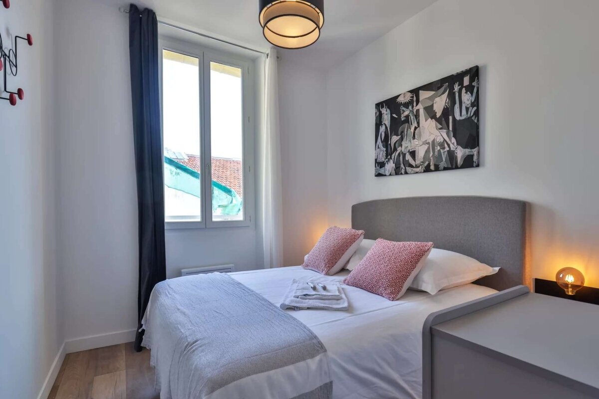 A modern, minimalist bedroom features a gray-headboard bed with pink cushions, white walls, a large window, abstract art, and a warm bedside lamp.