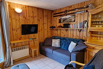 Cozy wood-paneled living room featuring a blue sofa, wall-mounted TV, decorative skis, and rustic wooden furniture.