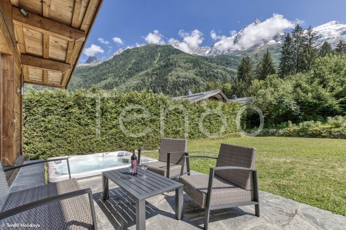 Chalet patio featuring a hot tub, outdoor seating with wine, and a lawn overlooking stunning, snow-capped mountains.