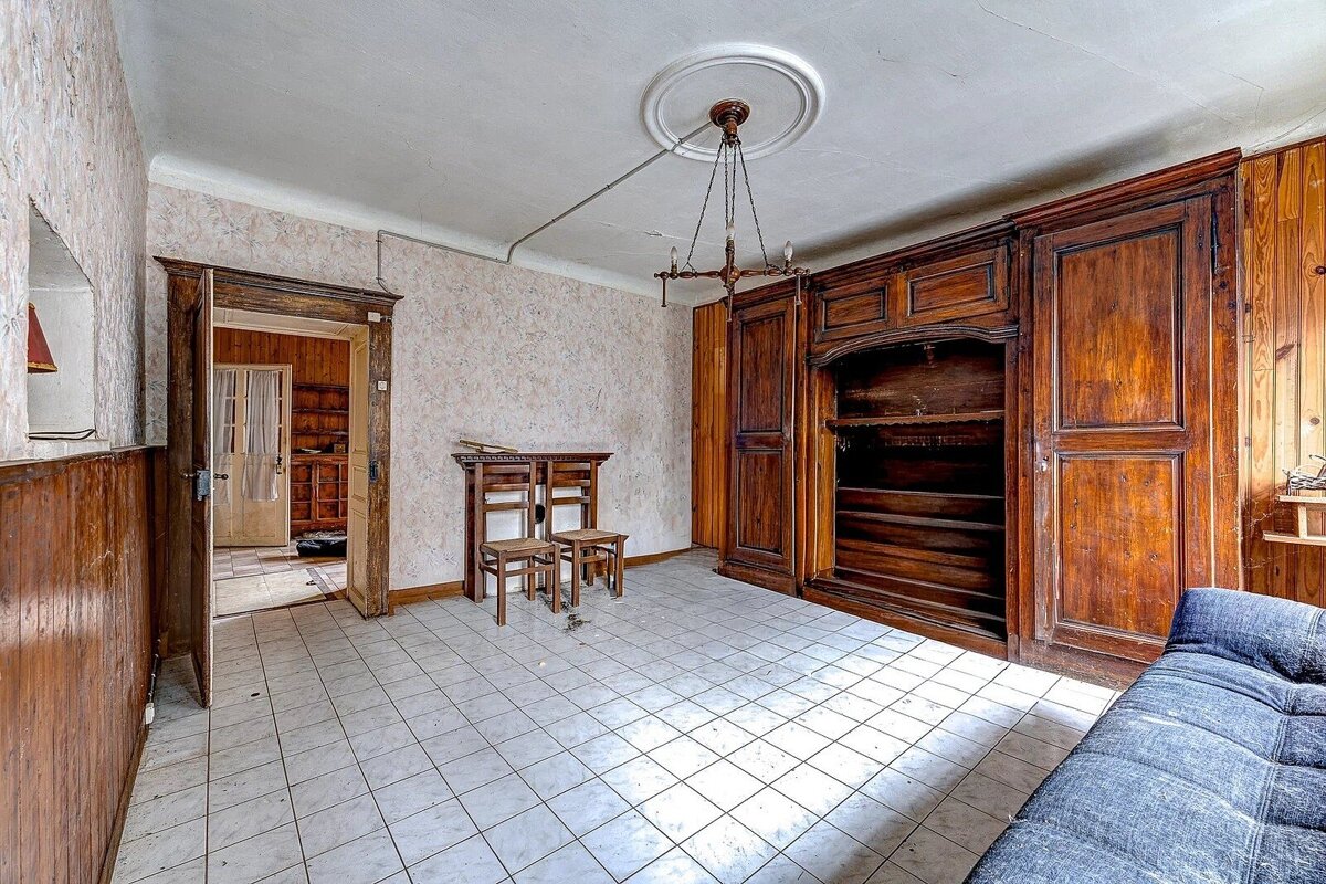 An old, worn room with large wooden furniture, tiled floor, patterned wallpaper, wood paneling, a chandelier, and a blue sofa.