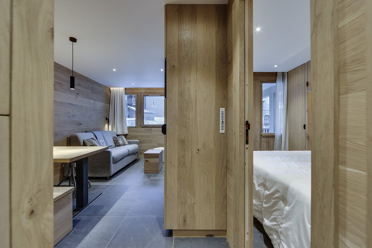 A modern, wood-paneled apartment interior showing a living area with a sofa and dining table, and a bedroom glimpsed through a sliding wooden partition.