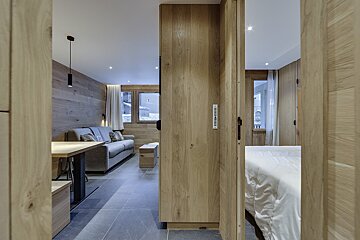A modern, wood-paneled apartment interior showing a living area with a sofa and dining table, and a bedroom glimpsed through a sliding wooden partition.