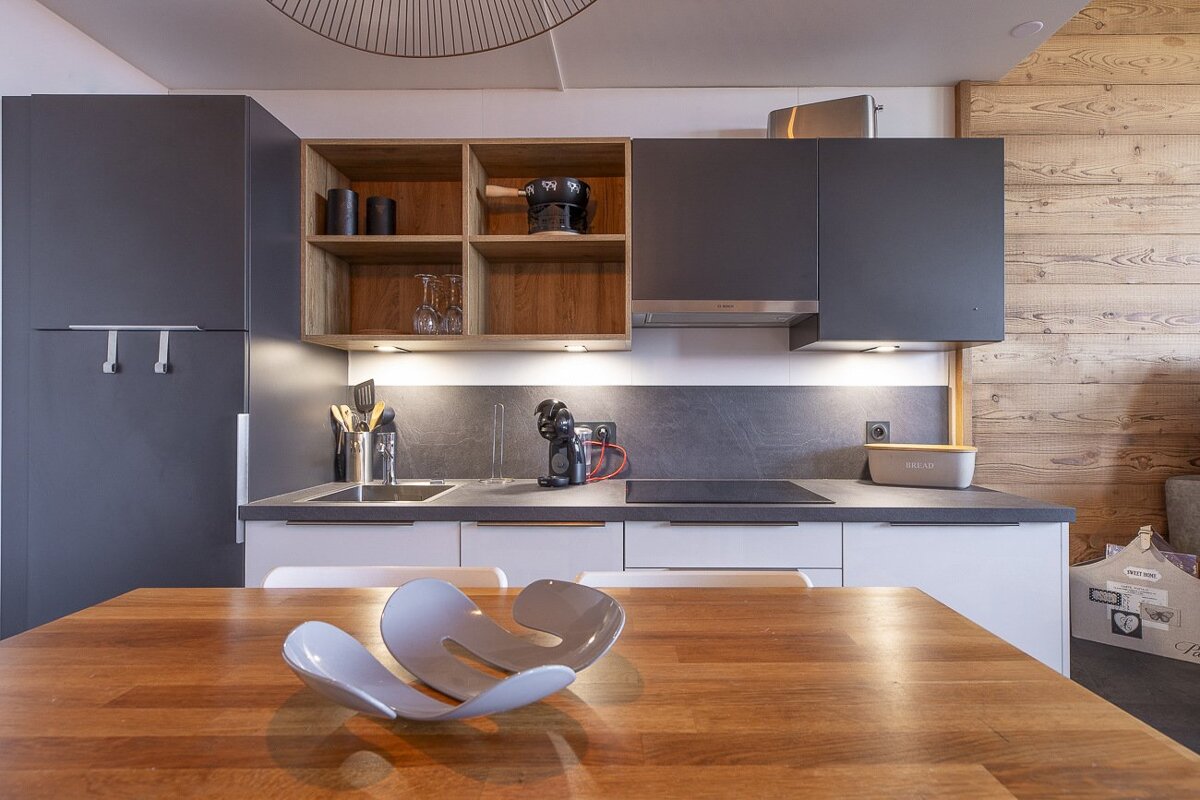 Modern kitchen with grey and wood cabinets, featuring a wooden dining table with a curved centerpiece in the foreground.
