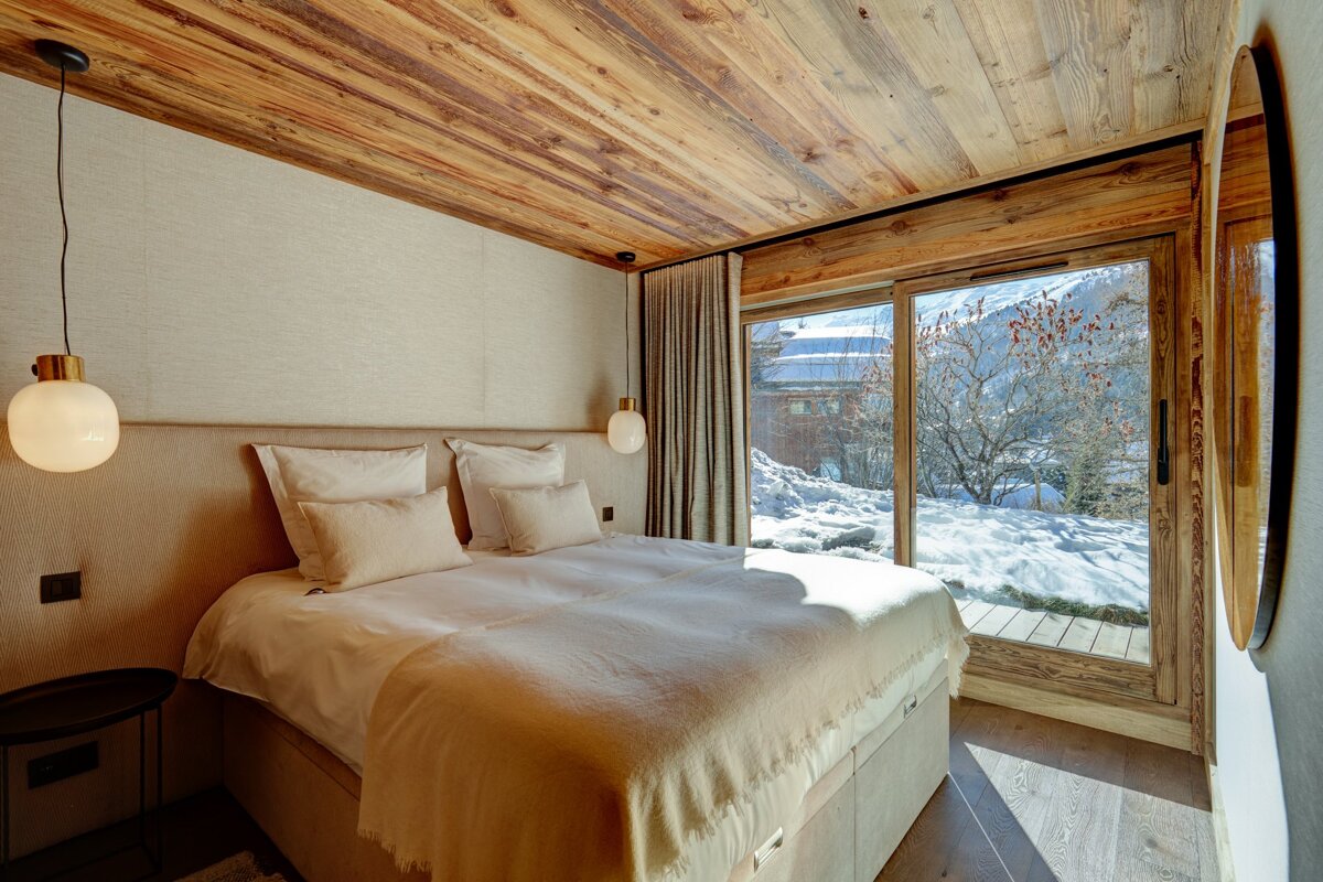 Chic bedroom with a warm wooden ceiling, neutral tones, and a large sliding door opening to a stunning snowy alpine view.