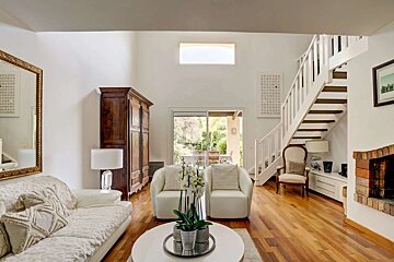 A spacious, bright living room featuring high ceilings, hardwood floors, white furniture, a wooden staircase, and a sliding door to an outdoor patio.