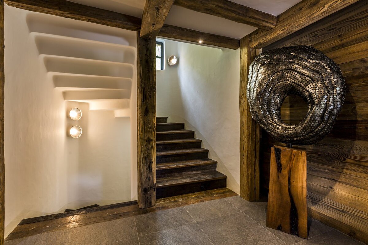 A rustic interior with exposed wooden beams, two staircases, white walls, and a striking metallic, textured ring sculpture.