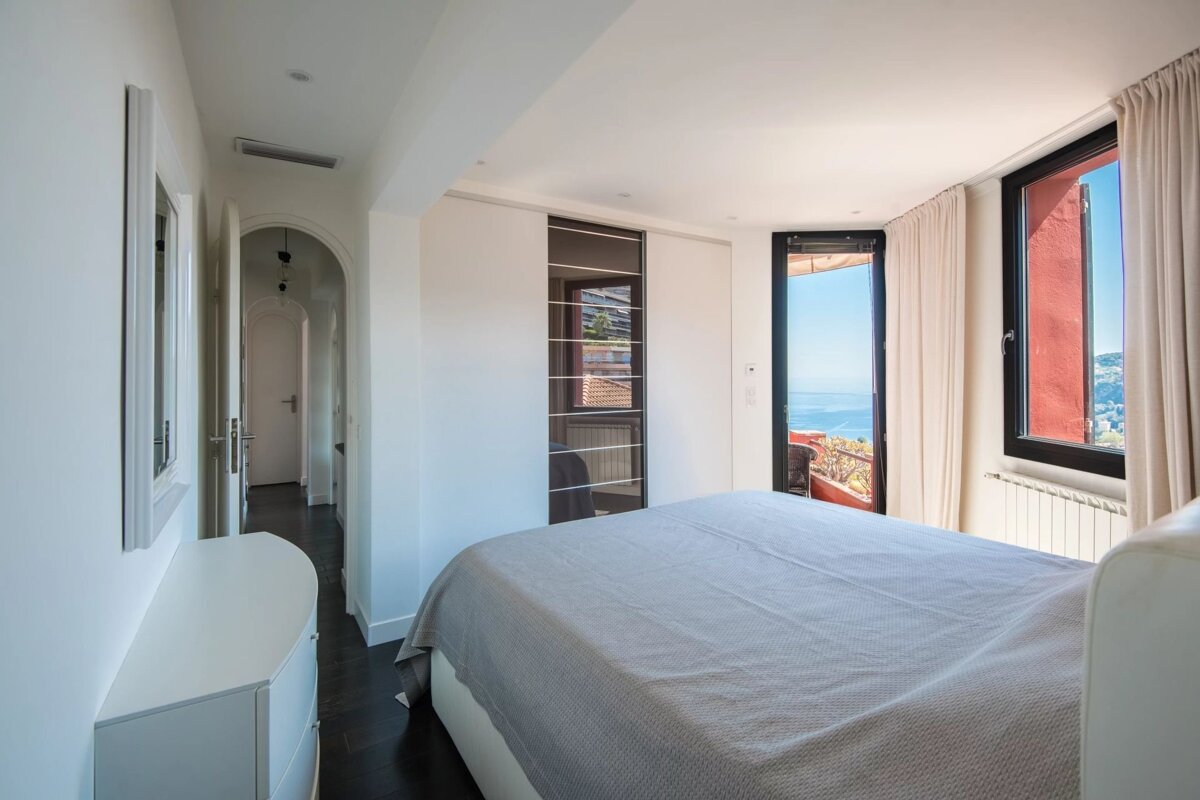 A bright, modern bedroom featuring a large bed, dark wood floors, and windows with a scenic ocean view.