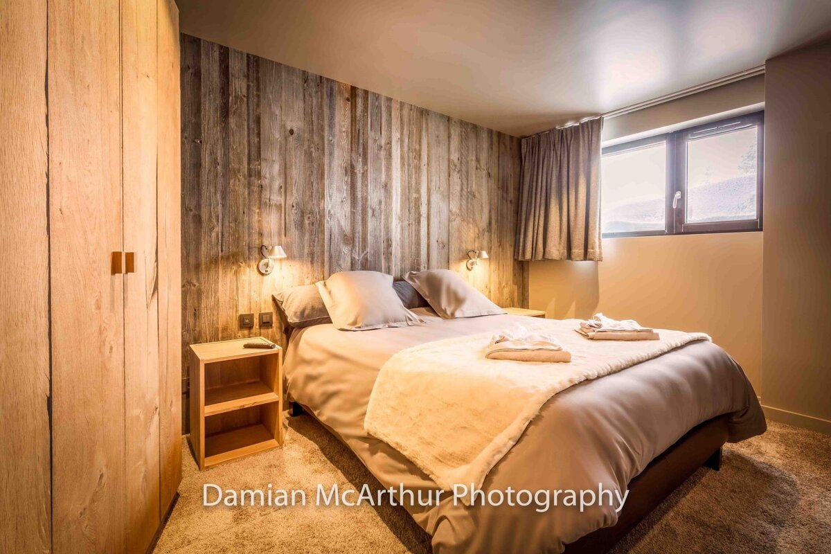 A cozy rustic bedroom featuring a wood-paneled wall, a double bed with beige linens, a wooden wardrobe, and a window.