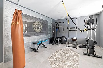 A modern home gym featuring a brown punching bag, elliptical, weight bench, pull-up station, and a decorative Medusa logo wall panel.