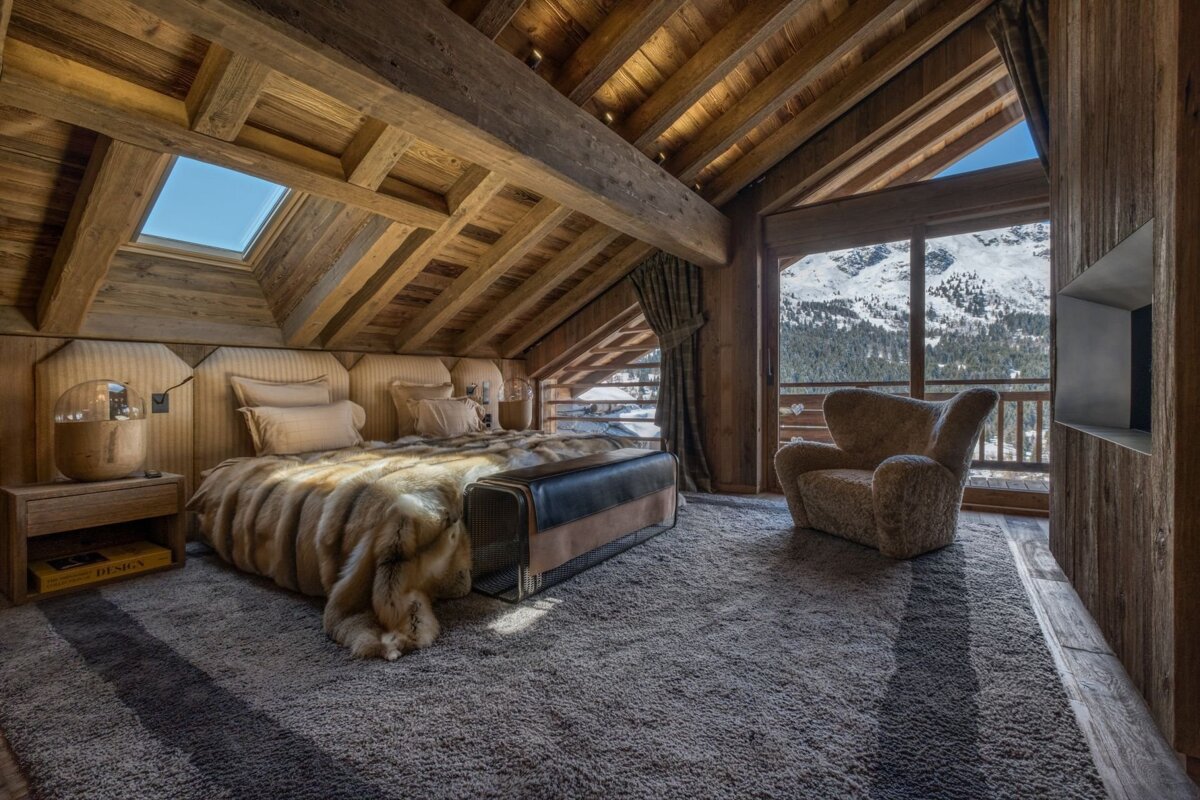 Rustic wooden bedroom featuring a fur blanket, skylight, and a large window with stunning snowy mountain views. A cozy armchair sits by.