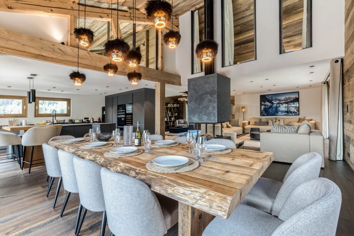 A luxurious chalet interior featuring a long wooden dining table, grey chairs, unique pendant lights, and a cozy living room.