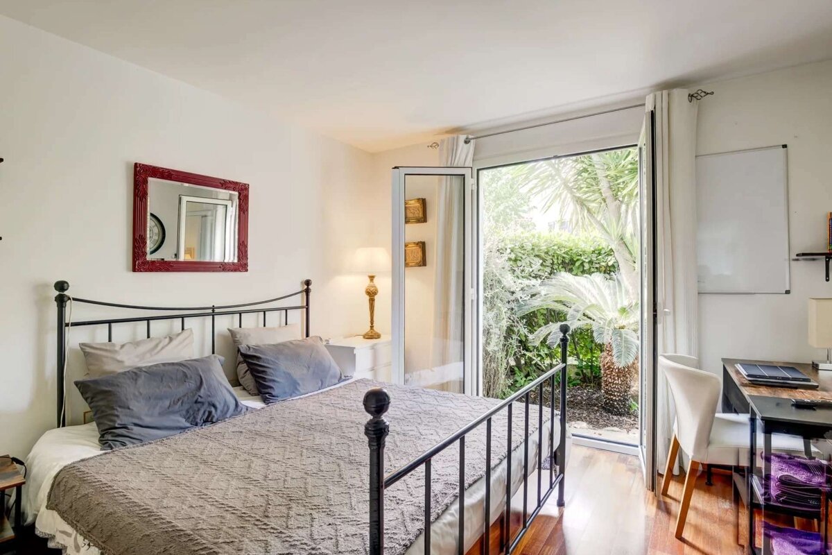 A bright bedroom with a wrought iron bed, red mirror, and desk. A glass door opens to a lush garden with palm trees, filling the room with light.