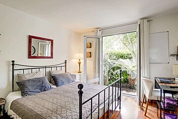 A bright bedroom with a wrought iron bed, red mirror, and desk. A glass door opens to a lush garden with palm trees, filling the room with light.