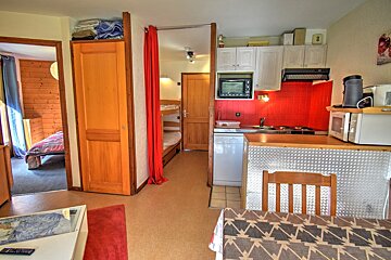 A compact apartment interior featuring a kitchen with red backsplash, a dining area, a bedroom with wood paneling, and bunk beds behind a red curtain.