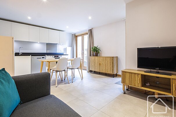 Modern open-plan living area with kitchen, dining table, gray sofa, TV, and balcony doors, featuring light colors and wood accents.
