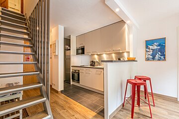 A modern kitchen with light cabinets and a bar with red stools, adjacent to a wooden staircase. Features mixed flooring and a mountain picture.