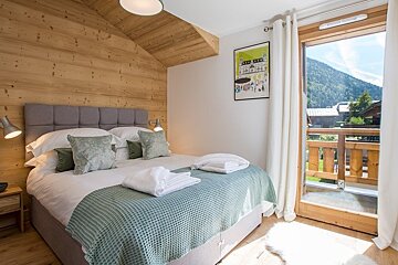 Cozy chalet bedroom with wood paneling, a large bed with green accents, and a glass door opening to a balcony with mountain views.