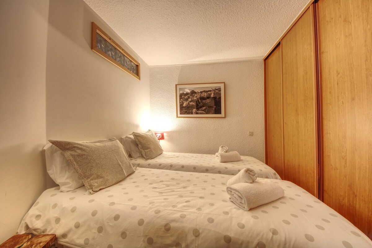 A cozy bedroom with two twin beds featuring white polka-dot duvets, white walls, a wooden wardrobe, and a framed picture of sheep.