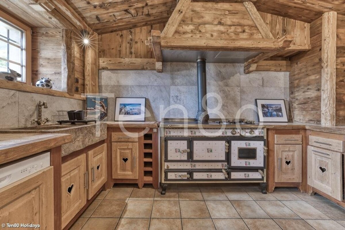 A rustic chalet kitchen with wooden paneling, heart-cutout cabinets, a stone sink, and a large vintage tiled stove under a wooden hood.