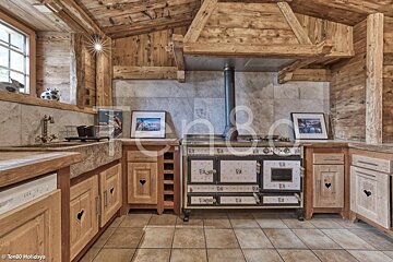 A rustic chalet kitchen with wooden paneling, heart-cutout cabinets, a stone sink, and a large vintage tiled stove under a wooden hood.