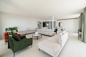 A spacious, bright, modern living room with sleek white floors, beige sofas, and a contrasting green velvet armchair.