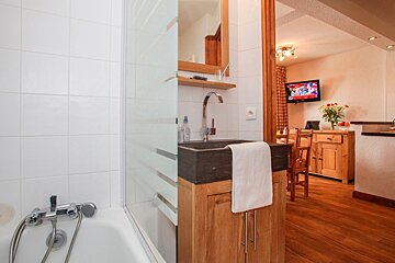 A bathroom with a tub and a modern stone sink flows into a cozy living area with a TV, dining space, and wooden furniture, showcasing an open-plan design.