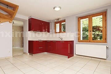 Modern kitchen with glossy red cabinets, white counter, tiled floor, and wooden windows. A wooden staircase is on the left.