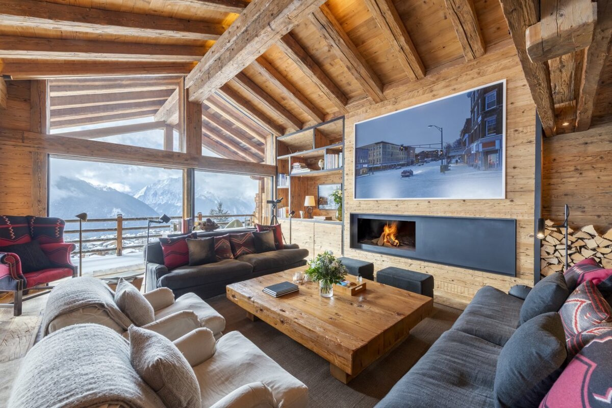Luxurious rustic chalet living room with a stunning mountain view, cozy fireplace, large sofas, and exposed wooden beams throughout.