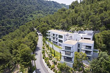 Modern white apartment buildings nestled on a lush, forested hillside next to a winding road.