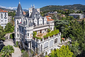 An ornate, multi-story mansion with spires, turrets, and terraces featuring outdoor seating, nestled amidst lush greenery and distant hills under a bright blue sky.